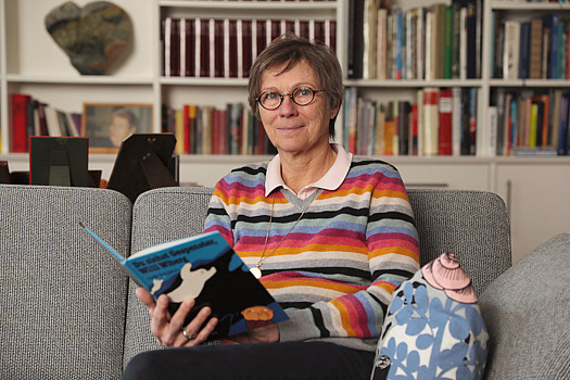 Nicola Ackermann-Aachner auf einem Sofa in einem Wohnzimmer mit großem Bücherschrank im Hintergrund.