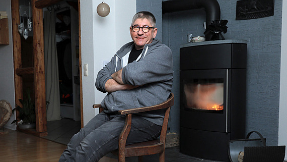 Dirk Wiezer sitzt in einem Raum auf einem Holzstuhl vor einem Kaminofen.