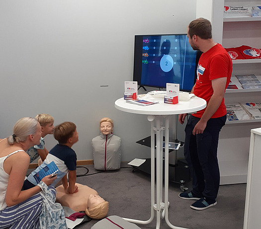 Zwei Kinder sowie eine Frau üben an einem Stand die Wiederbelebung anhand einer Dummy-Puppe und werden dabei von einer betreuenden Person angeleitet und digital unterstützt. einem Stand 