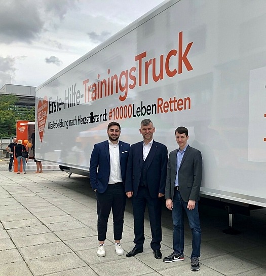 Drei junge Männer in Anzug vor dem "Erste Hilfe Trainings-Truck".